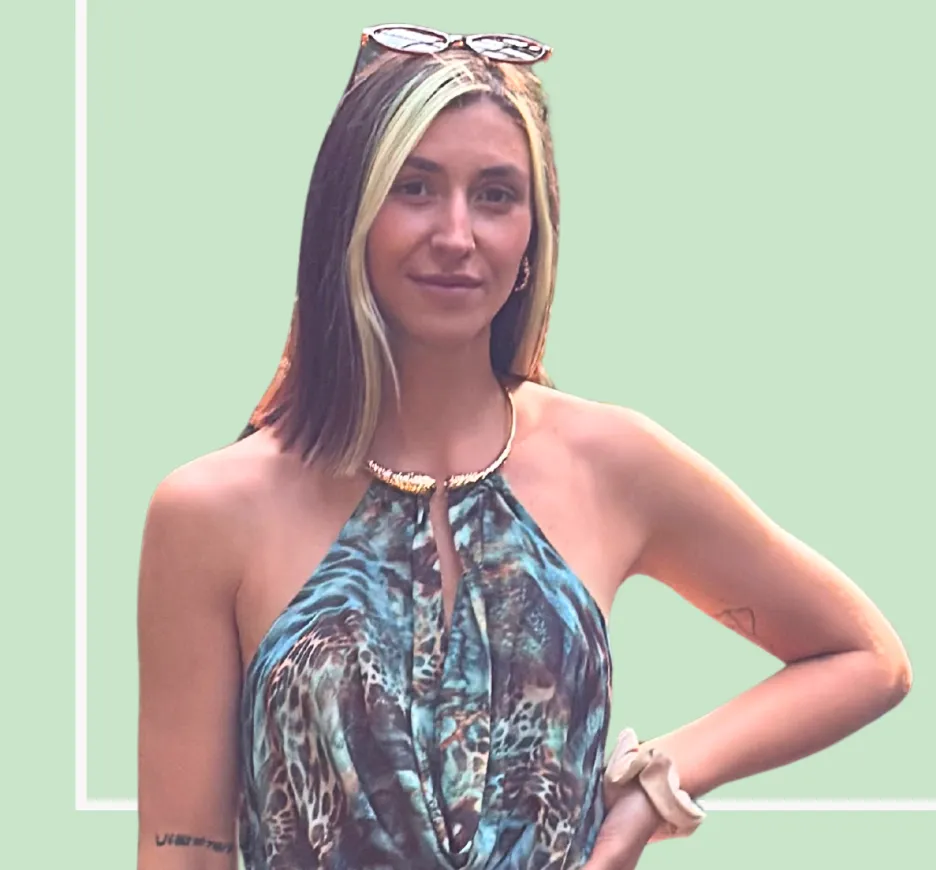 Sarah Althaus against a mint-green field: standing slightly turned, one hand on her hip, looking into the camera, wearing a patterned halterneck top, sunglasses pushed up into her hair and a gold necklace.